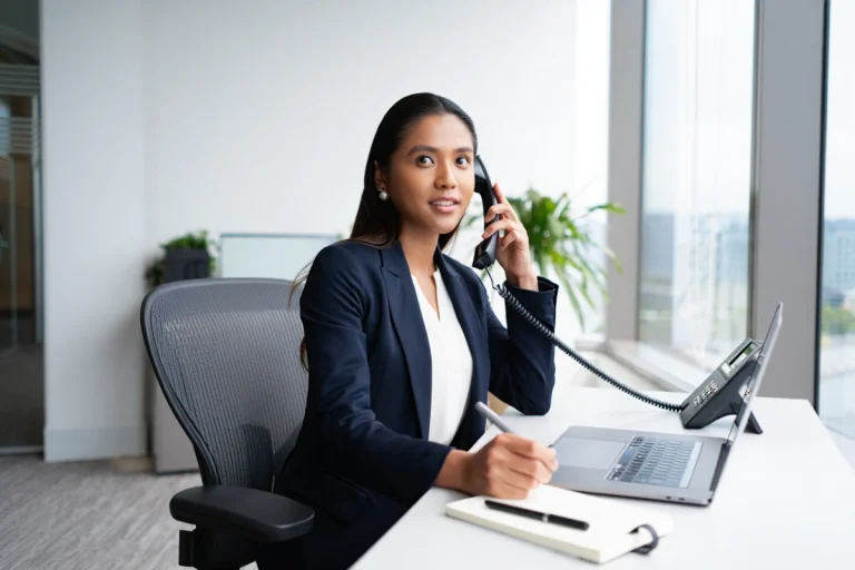 Secrétaire en costume sombre répondant à un appel téléphonique dans un bureau moderne, écrivant dans un carnet devant son ordinateur portable.